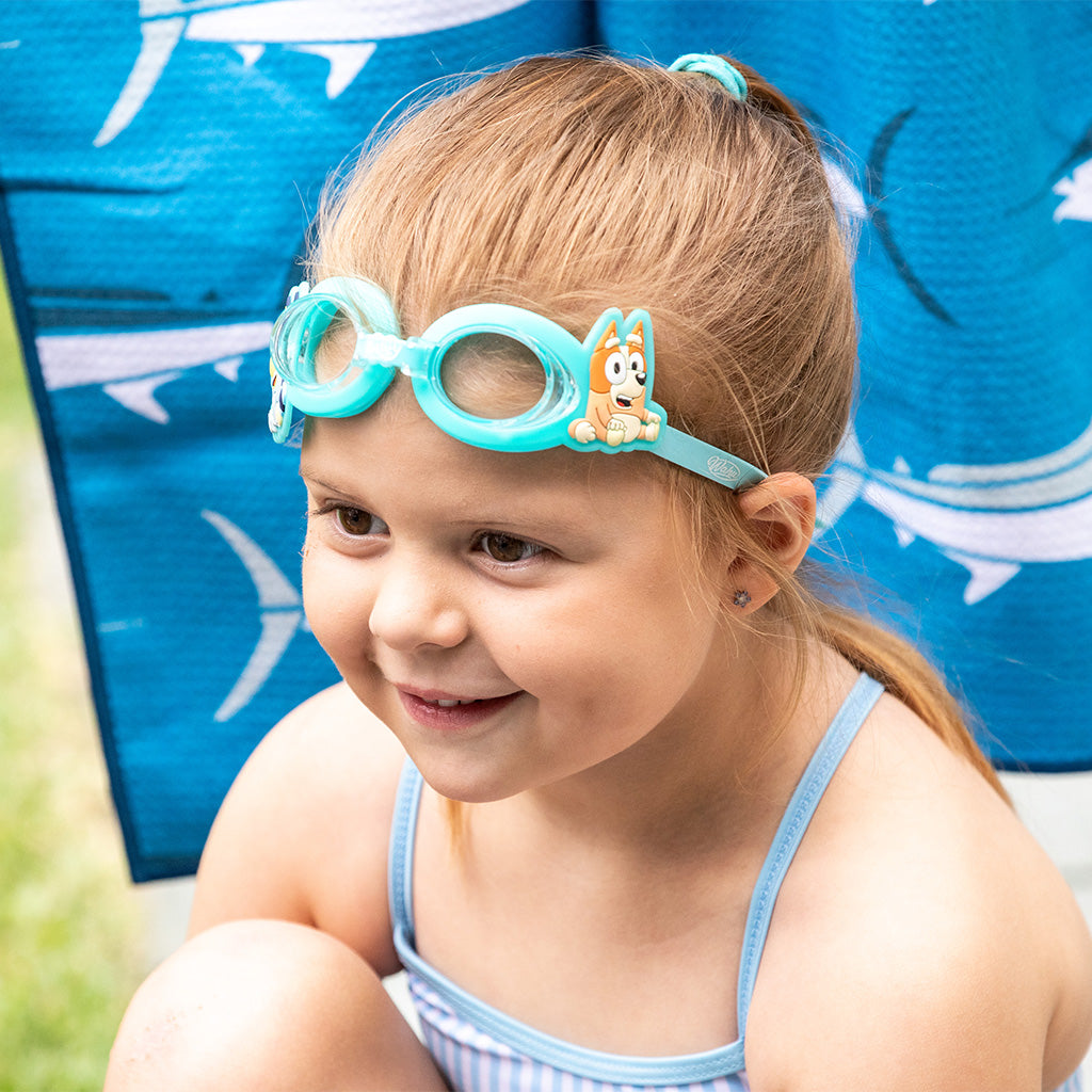 A young girl with blonde hair in a ponytail wearing turquoise Bluey swim goggles with Bluey and Bingo on the side, sitting outdoors in front of a blue towel.